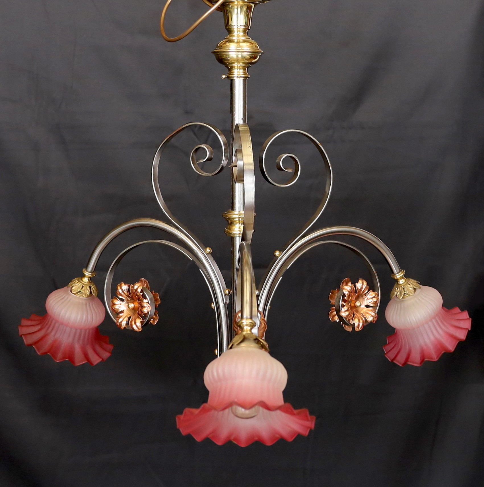 An English Arts & Crafts wrought iron brass and copper three light electrolier with pink tinted opaque glass shades, height 60cm. width 66cm